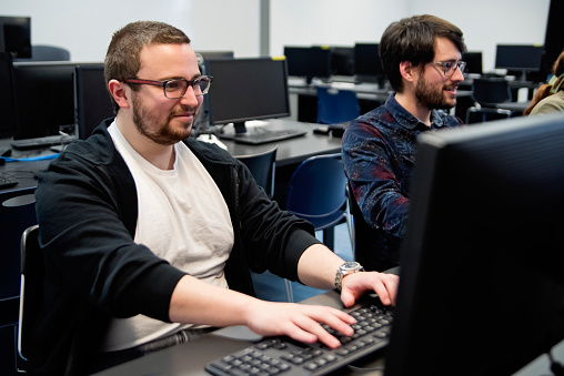 Two young adult male college students in programming classroom. They are sitting at desk using computers. Horizontal waist up indoors shot with copy space. This was taken in Quebec, Canada.