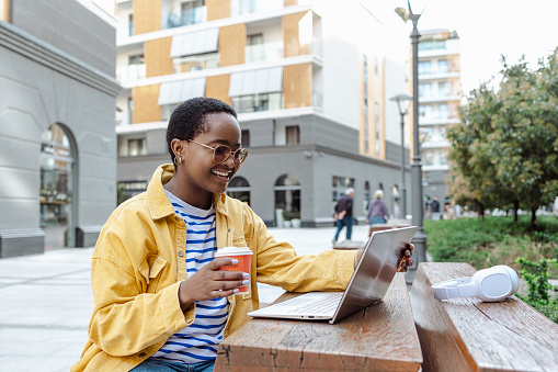 Portrait of a modern African American woman in casual clothes surfing the net on the laptop while drinking a coffee in the open-air cafeteria