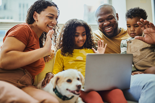 A young black family is sitting on a sofa in the living room with their dog while using their laptop computer for a video call while waving. Stock photo