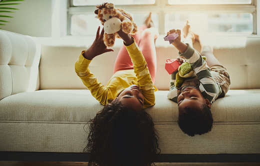 Brother and sister are lying upside down on the couch in the living room, holding up their toys, and talking with copy space. Stock photo