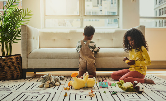 A brother kneeling with his back to the camera and a sister sitting on the rug playing with their toys, stock photo