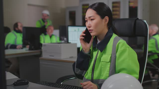 Asian woman technician in reflective uniform talk on phone and work on computer in control room