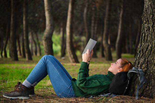 istock Redhead young woman reading a book lying under tree in forest. 1496499613