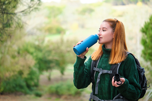 Smiley hiker redhead young woman drinking water from bottle in the mountain wearing a backpack. Trekking concept.