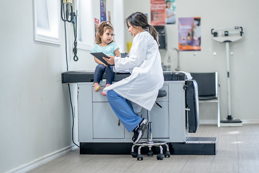 A sweet little girl sits up on an exam table during a doctors appointment.  She is dressed casually and her female doctor is seated on the table beside her in blue scrubs as they talk.