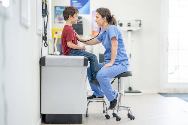 Talking a Close Listen A sweet young boy sits up on an exam table as his female nurse takes a close listen to his heart with her stethoscope. He is dressed casually and sitting still as she listens closely. red medical scrubs stock pictures, royalty-free photos & images