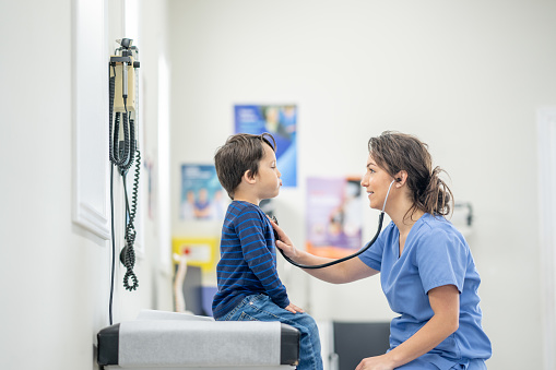 A sweet young boy sits up on an exam table as his female nurse takes a close listen to his heart with her stethoscope.  He is dressed casually and sitting still as she listens closely.