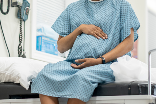 A young pregnant woman sit up on an exam table in her doctors office during a routine prenatal check-up.  She wearing a medical gown and holding her belly as she waits for her doctor.