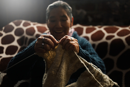 Abuela en la sala de casa feliz y concentrada, usando unos palitos de tejer y lana de oveja, concepto de vida cotidiana tradicional, tejedora tradicional, enfoque selectivo. Dia de los abuelos