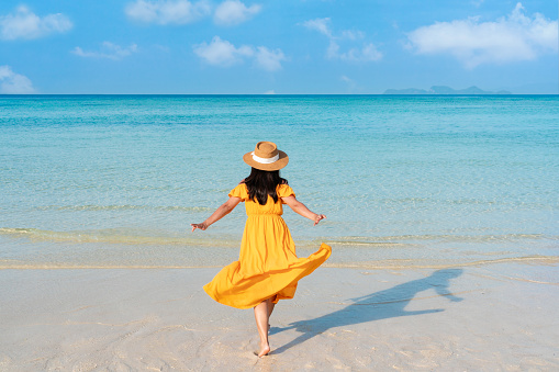 Happy young Asian woman in yellow dress run to the sea with white cloud on blue sky in travel vacation. Copy space