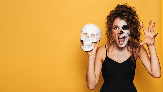 A young woman in a Halloween costume with makeup for the holiday on a yellow isolated background with a skull in her hands. The emotion on the face is fear-inducing. A place for your text or advertisement