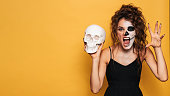 A young woman in a Halloween costume with makeup for the holiday on a yellow isolated background with a skull in her hands. The emotion on the face is fear-inducing. A place for your text or advertisement.