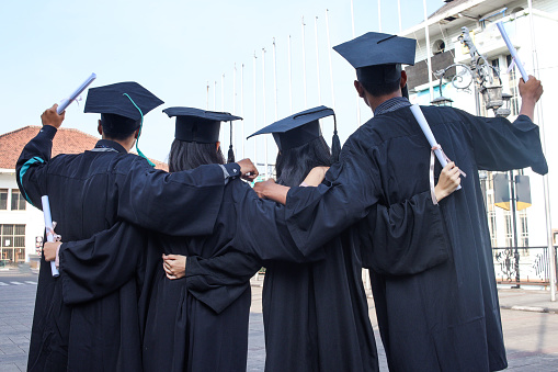 Rear view of a group of high school graduates wearing caps and gowns, with their arms around each other, on white background, couple grabbing each others rear end.