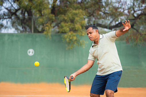Indian senior man busy playing tennis or servicing by hitting ball with racquet at court - concept of active healthy lifestyles,fitness and sports hobbies
