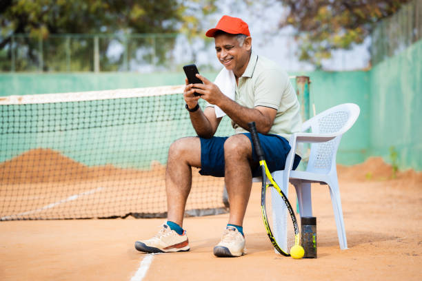 istock Happy indain senior man at tennis court busy using mobile phone during break while playing game - concept of technology, social media and relaxation 1496560499
