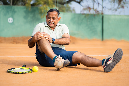 Indian fallen injured senior man suffering from knee pain while playing tennis at court - concept of emergency, painful and inflammation