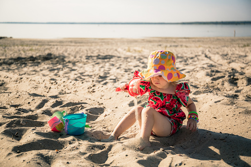 A little cute 3 year old girl in red dress and panama hat sits on the beach and plays in the sand with plastic toys: a bucket, molds and a shovel. Summertime rest. Family vacation. Traveler. Close-up.