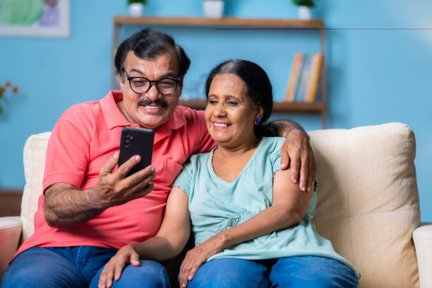 istock Happy Indian senior couple using on mobile phone while sitting on sofa at home - concept of social media and retirement lifestyles and internet connection 1496572318