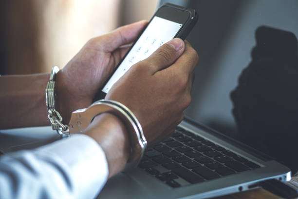 istock Close up hands of hacker holding mobile phone with handcuffs above notebook keyboard, punishment for cybercrime, business concept. 1496573758