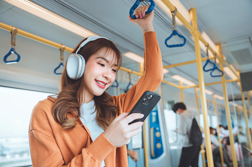 Happy young Asian woman passenger listening music via smart mobile phone in a train, Smile female wearing wireless headphones while moving in the tram, lifestyle, transportation.