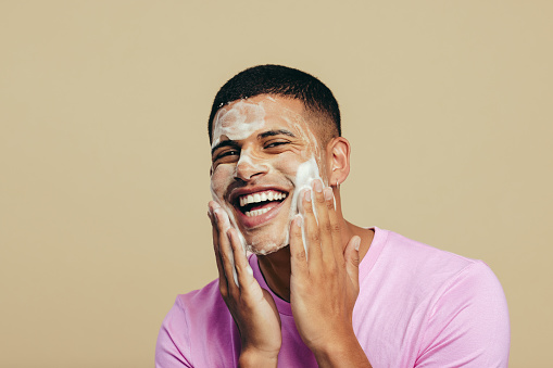 Portrait of a handsome young man indulging in his grooming and self-care routine as he applies face wash in a studio. Youthful man smiling happily while massaging his skin with a facial cleanser.
