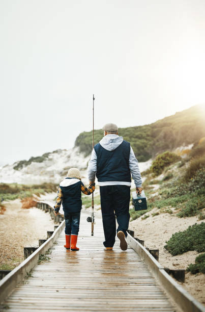 istock Fishing, child and grandfather walking on beach pier with tools from back, bonding time on weekend with mockup. Nature, family and old man with boy holding hands and learning catch fish in Greece. 1496581128