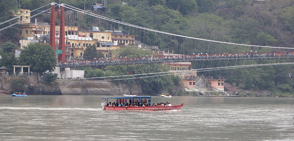 The boat goes along the river towards the bridge. \nRam Jhula bridge across the Ganges River in Rishikesh. Ram Jhula is an iron suspension bridge situated at Muni Ki Reti in Rishikesh in the Indian state of Uttarakhand. Built in 1986, over river Ganges to cross the river. Rishikesh. Uttarakhand. India. Asia. 06-18-2019