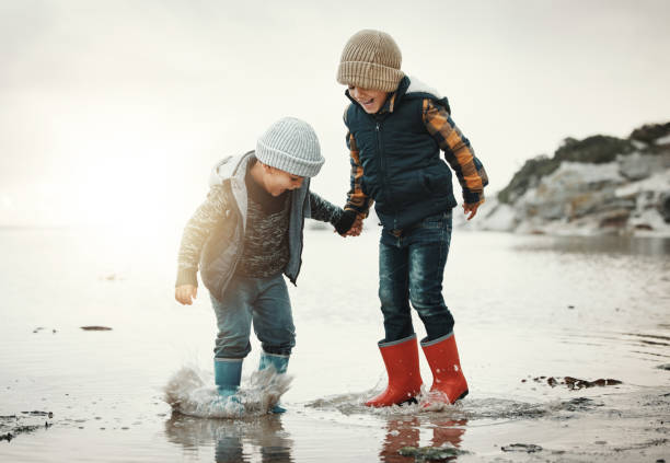 ビーチ、ブーツ、水の中の子供たちが一緒に手をつないで、笑顔で波の中で遊ぶ。楽しい、休日と兄弟、日没時の海の休暇の幸せな男の子、夕方の海の散歩でジャンプして水しぶき。 - fun walk ストックフォトと画像