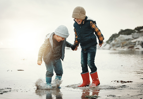 Beach, boots and children in water together, holding hands and playing in waves with smile. Fun, holiday and brothers, happy boys on ocean vacation at sunset, jumping and splash on evening sea walk.
