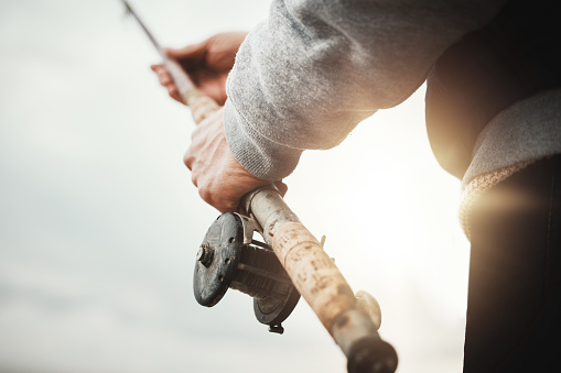 Hands, fishing and rod with a man in nature, enjoying a weekend trip his hobby or pastime at sunset. Morning, flare and low angle with a male fisherman casting a line outdoor in the wilderness