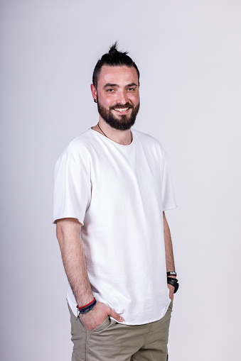 Portrait of charming young man standing against a white background, hands in pockets, smiling at camera with confidence.