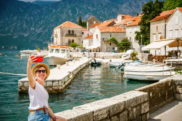 Tourist woman making a selfie sitting on the embankment of the beatiful small town Perast, Kotor Bay, Montenegro