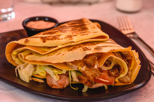 Big Pancake filled with chicken with cheese, bacon, tomato, lettuce, sauce on a black plate in greek tavern. Thin pancakes, crepes. Mediterranean cuisine. Selective soft focus