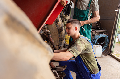 In a display of teamwork, a mature man and his son collaborate in their workshop to repair a tractor.