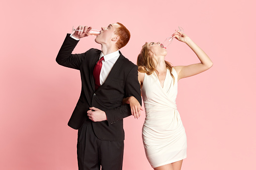 Young man and woman, couple emotionally drinking champagne after getting married against pink studio background. Expressive look. Concept of family, relationship, youth, emotions, fun. Ad