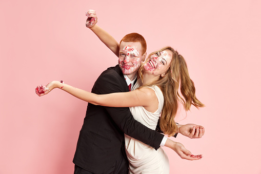 Portrait of happy young married couple, man and woman celebrating marriage, eating cake with dirty faces against pink studio background. Concept of family, relationship, youth, emotions, fun. Ad
