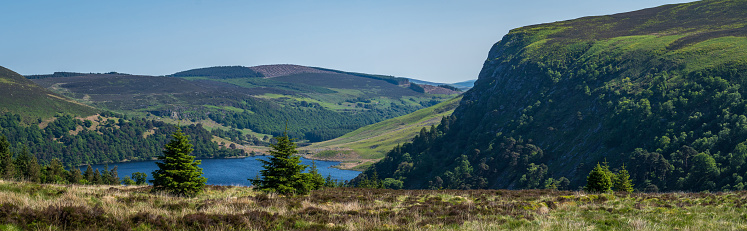 Panoramic wide view of the cliff face running into Lough Tay in the Wicklow mountains, on a bright sunny summer day with blue sky. The area is a popular tourist attraction with spectacular scenery