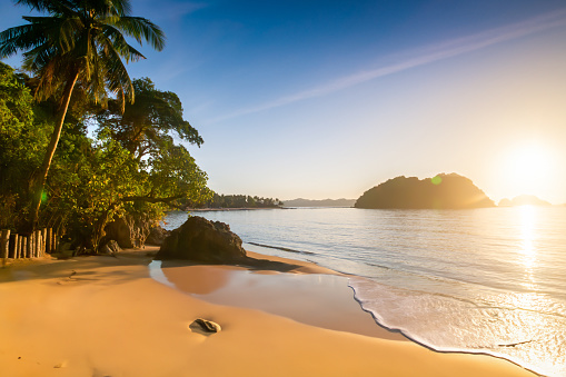 Sunset scene at a beach near El Nido in the Philippines. The calm sea reflects the warm glow of the setting sun. The sandy shore appears clean and inviting. On the left side of the frame, palm trees and tropical plants are visible, adding a touch of greenery to the landscape. In the distance, multiple islands can be seen. The sky above is clear, with minimal cloud cover.