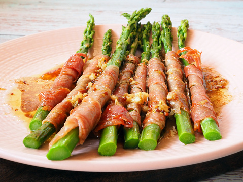 Сlose-up showcasing a hot appetizer of green asparagus delicately wrapped in savory ham or bacon slices. The dish is beautifully enhanced with a drizzle of sweet and sour garlic sauce.