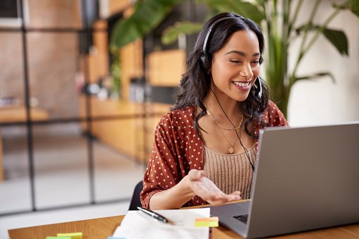 Smiling successful business woman with headphones talking while attending a meeting online using laptop. Latin woman using computer with headphone and microphone in a online meeting. Hispanic businesswoman watch webinar and listen online course.