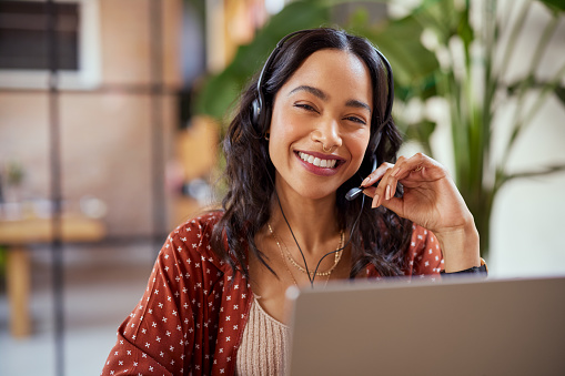 Smiling businesswoman working in a creative office. Smiling friendly woman working as call center agent for online support. Mixed race girl in video call with headphones looking at camera with copy space.