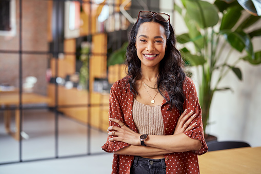 Portrait of smiling young multiethnic woman looking at camera with crossed arms. Successful latin business woman standing in modern office with copy space. Young university hispanic girl with pleasant smile and folded arms.