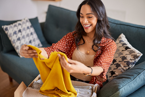 Happy young latin woman checking clothes sitting on sofa at home. Cheerful shopaholic hispanic woman holding new clothes. Smiling multuethnic girl unpacking parcel box with clothes after shopping online.