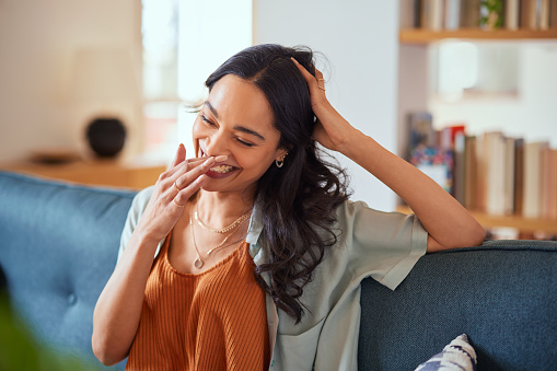 Happy young latin woman sitting on sofa at home and laughing. Happy multiethnic carefree woman in casual clothing relaxing at home while laughing with hand near mouth. Close up face of cheerful girl sitting on couch and laughing while thinking of something funny.