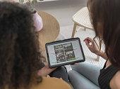 Rear view of a young female couple sitting in new home, searching and watching home decorating style on tablet. Happy young adult women couple sitting on couch in new house, using digital tablet, trying to find reference for home decorations online.