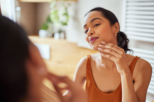 Pretty young woman touching and examining her face skin while looking in mirror in the morning. Beautiful latin woman enjoying hydrated skin in her bathroom. Happy self loving girl enjoying her natural beauty complexion, during morning beauty routine.