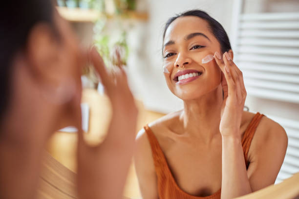 smiling mixed race young woman applying moisturizer on her face in bathroom - attractive female indoors lifestyles clean imagens e fotografias de stock