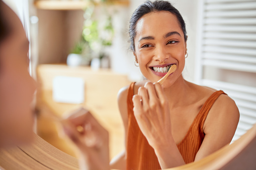 Smiling young woman brushing teeth in bathroom. Happy girl looking in mirror while using ecological toothbrush with whitening toothpaste to clean her teeth. Multiethnic girl cleaning teeth in the morning time.