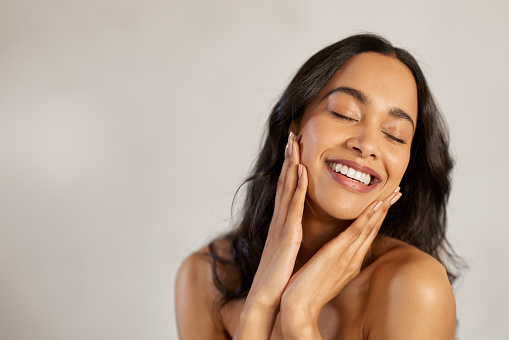 Portrait of beautiful latin woman touching her clean and healthy face against grey background. Smiling hispanic woman with natural makeup feeling healthy skin with eyes closed. Attractive multiethnic young woman pampering with perfect skin isolated with copy space.