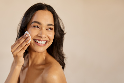Beautiful young multiethnic woman with bare shoulder removing make up with cotton pad on cheek isolated against beige background. Smiling hispanic girl applying dry powder using cosmetic cushion on skin. Skincare and beauty routine concept.